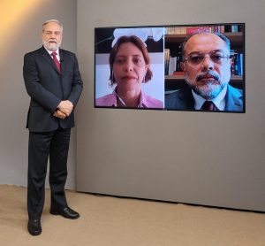 Foto do estúdio do programa com o apresentador posando ao lado do telão onde se vê as pessoas entrevistadas por videoconferência.