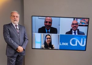 Foto do estúdio do programa com o apresentador posando ao lado do telão onde se vê as pessoas entrevistadas por videoconferência.