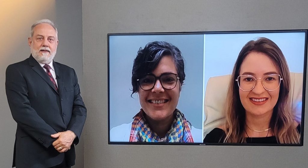 Foto do estúdio do programa com o apresentador posando ao lado do telão onde se vê as pessoas entrevistadas por videoconferência.