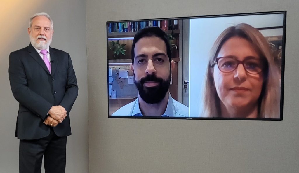 Foto do estúdio do programa com o apresentador posando ao lado do telão onde se vê as pessoas entrevistadas por videoconferência.