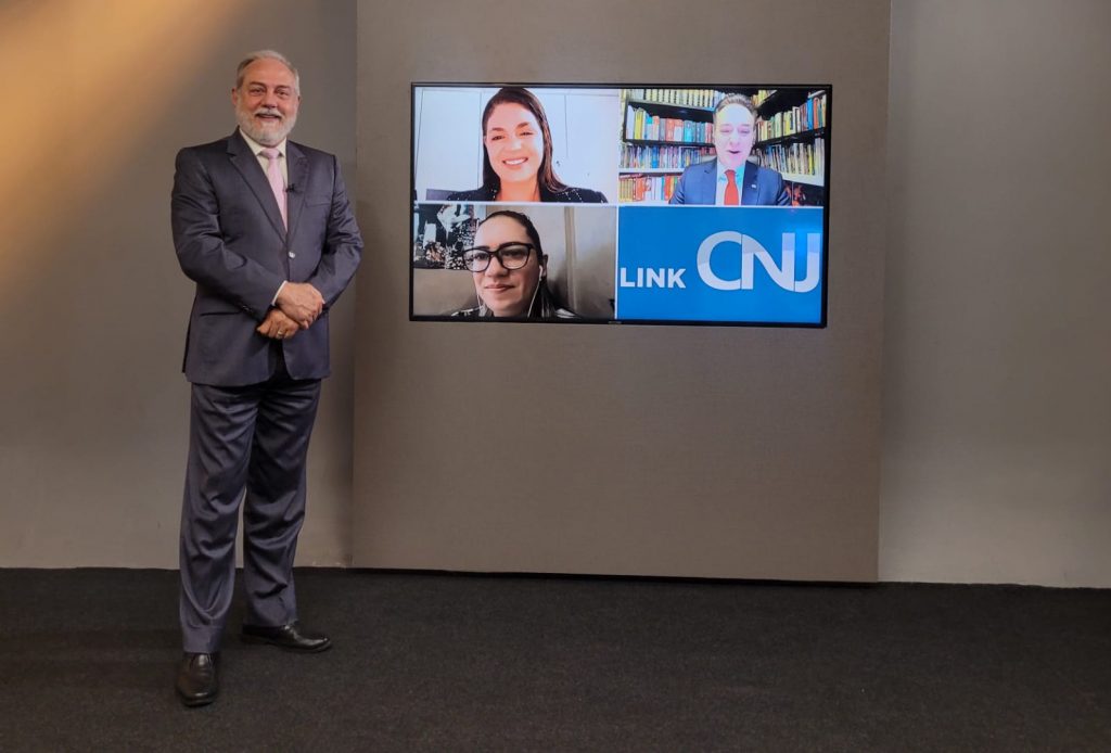 Foto do estúdio do programa com o apresentador posando ao lado do telão onde se vê as pessoas entrevistadas por videoconferência.
