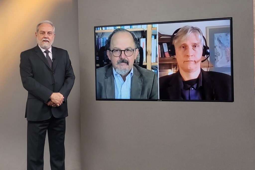 Foto do estúdio do programa com o apresentador posando ao lado do telão onde se vê as pessoas entrevistadas por videoconferência.