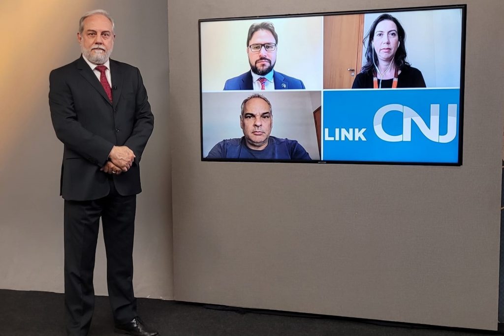 Foto do estúdio do programa com o apresentador posando ao lado do telão onde se vê as pessoas entrevistadas por videoconferência.