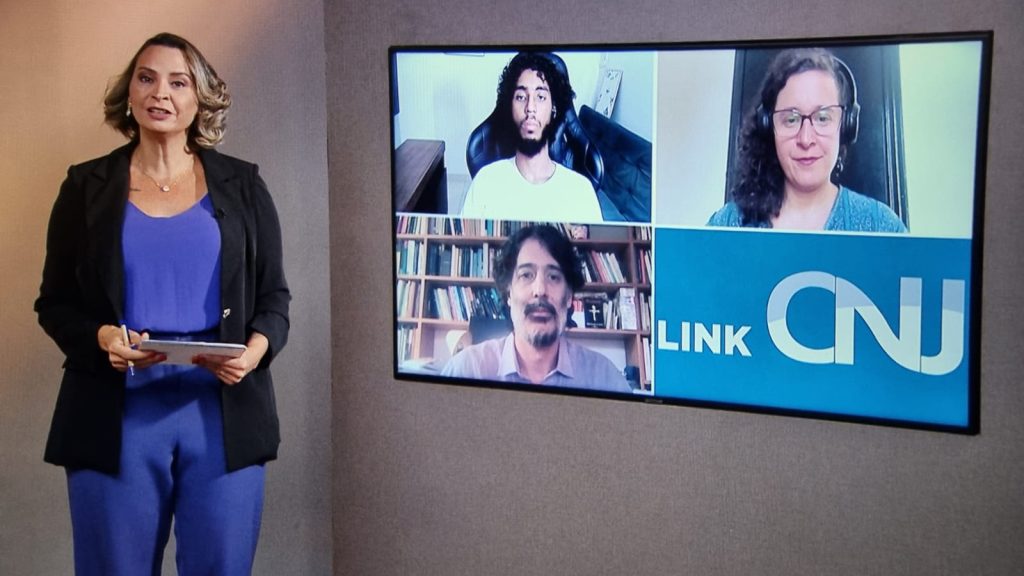 Foto do estúdio do programa com o apresentador posando ao lado do telão onde se vê as pessoas entrevistadas por videoconferência.