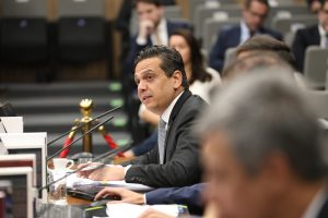 Fotografia em plano médio; Conselheiro do Conselho Nacional de Justiça, Marcos Vinícius Jardim. Sentado sobre a mesa plenária, vestido formalmente, terno de tom escuro.