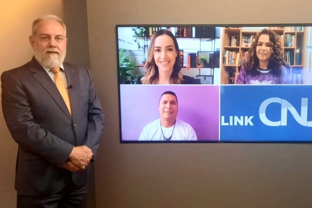 Foto do estúdio do programa com o apresentador posando ao lado do telão onde se vê as pessoas entrevistadas por videoconferência.