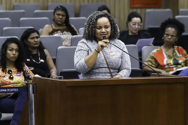 Imagem da participação da Senhora Antônia Cariongo - Liderança Quilombola e Defensora dos Direitos Humanos, durante a Audiência Pública no auditório do Conselho Nacional de Justiça. Ao centro em pé segurando o microfone com a mão direita, vestida formalmente, blusa em tom acinzentado. Ao fundo, desfocado há algumas cadeiras e mulheres sentadas.