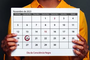 Imagem em plano detalhe, sobre fundo cinza uma pessoa vestindo uma blusa amarela com um calendário em mãos, marcando o dia vinte de novembro - Dia da Consciência Negra.