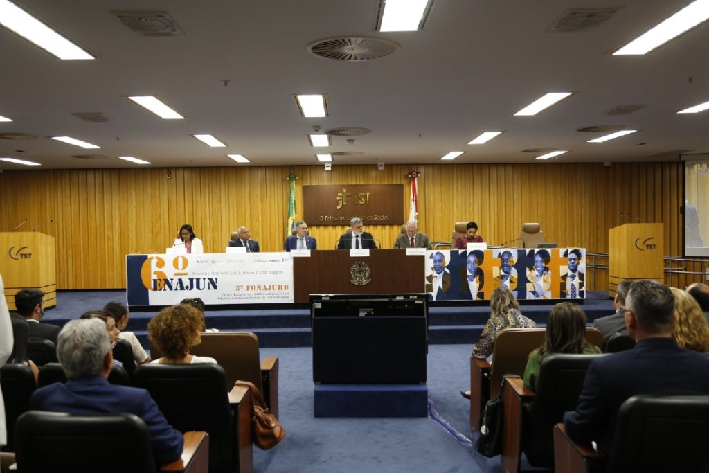 Imagem em plano geral do auditório do TST Tribunal Superior do Trabalho. Ao fundo parede em tom amadeirado, compondo a sala ao centro mesa composta por seis pessoas, quatro homens e duas mulheres, dentre eles o ministro Luís Roberto Barroso, todos vestidos formalmente. Na plateia sentados, homens e mulheres.