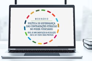 Em plano detalhe; Notebook em cima de uma mesa, sobre a tela em fundo branco dentro de um círculo pontilhado colorido, o texto; WEBINÁRIO - Política de Governança das Contratações Públicas no Poder Judiciário: Nível de Implementação da Resolução CNJ N.347/2020 e Boas Práticas.