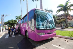 Foto em plano geral, em destaque um ônibus rosa parado e um pequeno grupo de mulheres concentradas em sua porta.