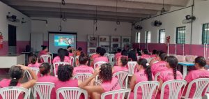 Imagem em plano geral; Sala nas cores rosa e branco, na lateral direita cinco janelas gradeadas e ventiladores de parede nas laterais superiores. Ao centro, mulheres sentadas em cadeiras de plástico branco, vestidas com blusas de tom rosa, na parte de trás da blusa está a palavra: INTERNA.