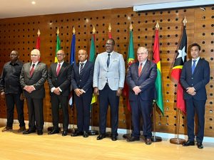 Sete senhores enfileirados em pé, sobre parede em tom amadeirado, vestidos formalmente, dentre eles o vice presidente do CNJ, ministro Edson Fachin. Atrás, oito bandeiras dos Países de Língua Portuguesa.
