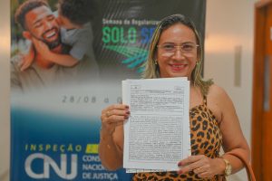 Fotografia em plano médio: mulher com um documento em mãos, cabelos soltos na altura do ombros, usando óculos de grau e blusa com estampa de onça. Ao fundo um banner com o texto: Semana da Regularização Fundiária Solo Seguro.