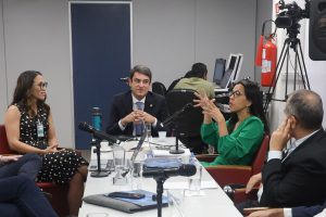 Sala de paredes brancas e porta de tom azul, composta por uma mesa ao centro e seis pessoas dialogando, entre eles homens e mulheres. Ao fundo duas pessoas sentadas aparentemente usando computadores.