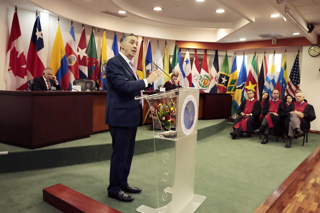 Fotografia da sala da sede da corte Interamericana de direitos humanos, composta pelas bandeiras dos países representantes, na lateral direita estão presentes, juristas, advogados e outros, todos sentados e vestidos formalmente. A sala possui o piso aveludado em tom de verde, paredes brancas e ao centro em pé está o ministro Luís Roberto Barroso em discurso, vestido formalmente.