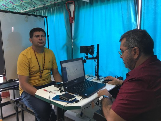 A imagem é um registro fotográfico horizontal, feito no ônibus do TRE-CE, na ação itinerante na Caucaia. Está presente, do lado esquerdo, um eleitor de blusa amarela e calça jeans. Ao centro, há uma mesa branca com um notebook e uma câmera. Do lado direito, o servidor do TRE-CE com uma blusa vermelha.