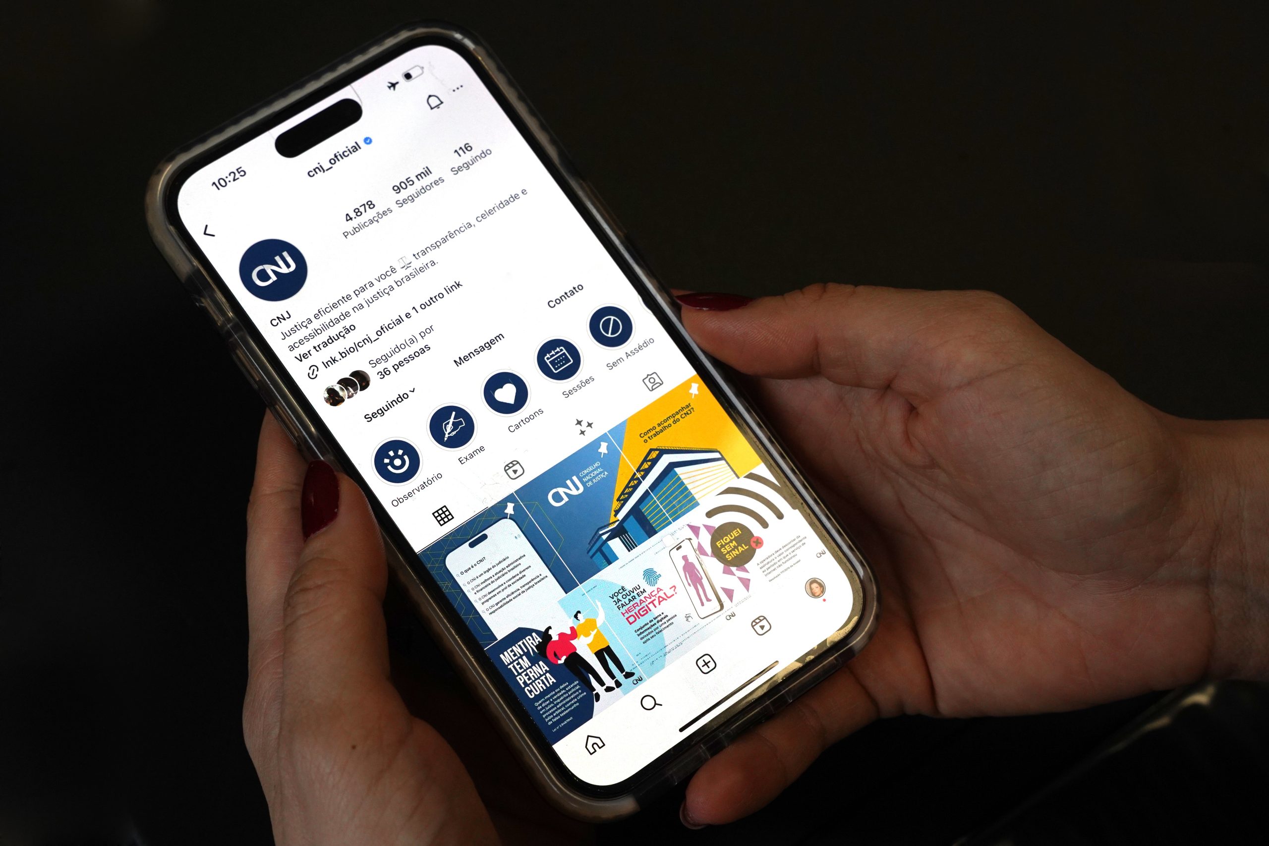 Em plano detalhe, sobre fundo escuro, mãos segurando um celular, na tela a nova atualização da identidade visual das redes sociais do Conselho Nacional de Justiça.