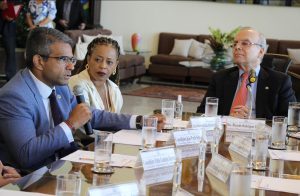 Imagem em plano médio composta por grupo de três pessoas sentadas, dentre eles; Conselheiro João Paulo Schoucair, vestido formalmente, usando óculos de grau e segurando um microfone na mão direita. Ao centro, Secretária-Geral do CNJ, Adriana Cruz, vestida em um terno formal de tom branco, ao lado direito o desembargador Ricardo Rodrigues Cardozo, vestido formalmente. Sobre a mesa, copos com agua, placas com nomes, papeis e canetas.