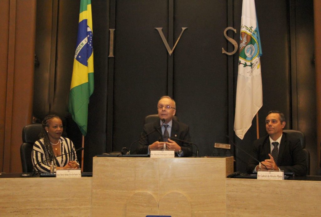 Fotografia em plano médio da sala do Tribunal de Justiça do Rio de janeiro, composta por um fundo preto ao centro acima um letreiro com as letras IVS, nas laterais duas bandeiras. Abaixo centralizado dois representantes do Conselho Nacional de Justiça e um do Tribunal de Justiça do Rio Janeiro, sentados e vestidos formalmente.