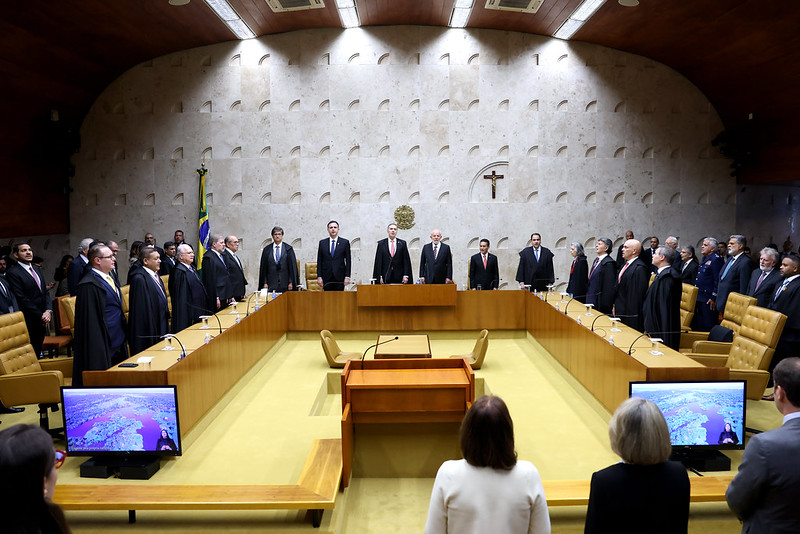 Fotografia em plano geral; Plenário do Supremo Tribunal Federal, composta por uma mesa em formato de U ao centro, ao fundo parede branca com brasão do STF, bandeira do Brasil e um crucifixo. Na mesa estão presentes os ministros, desembargadores e o presidente da República, todos em pé e vestidos formalmente.