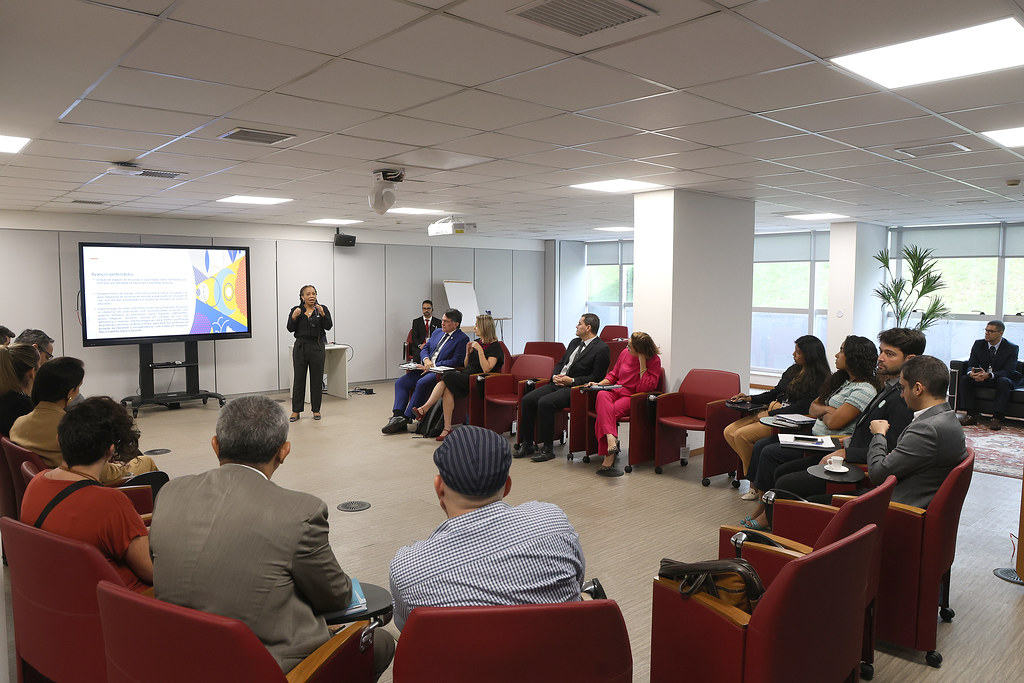 Fotografia em plano geral de uma sala de reuniões, ao centro um círculo formado por cadeiras vermelhas e pessoas sentadas. Ao fundo uma tela de transmissão e uma mulher, aparentemente mediadora da oficina.