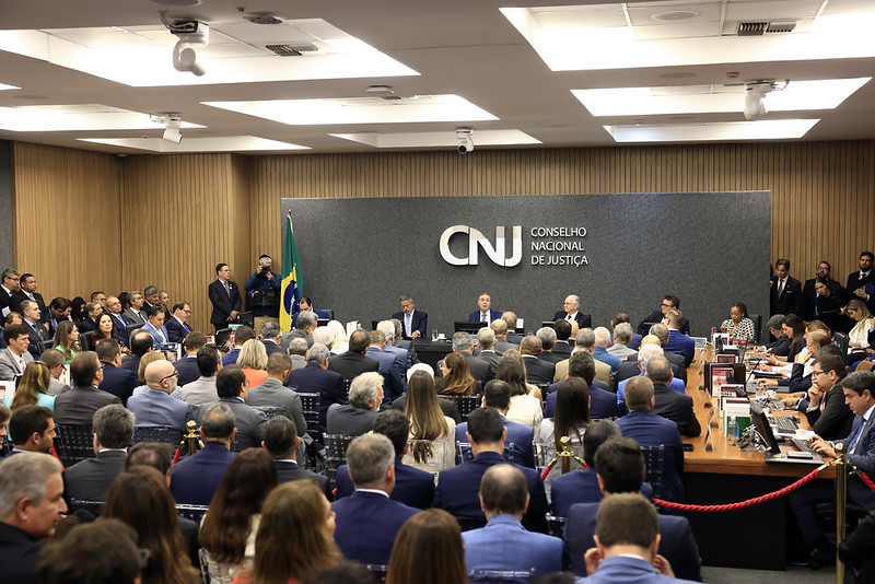 Fotografia em plano geral do plenário do Conselho Nacional de Justiça, durante a solenidade de posse dos conselheiros e conselheiras. Ao fundo a logo do CNJ sobre um painel de tom acinzentado, ao centro uma mesa em formato de “U”, composta pelo ministro Luís Roberto Barroso e os conselheiros (as), e em primeiro plano a plateia presente durante a cerimônia, todos sentados e vestidos formalmente.