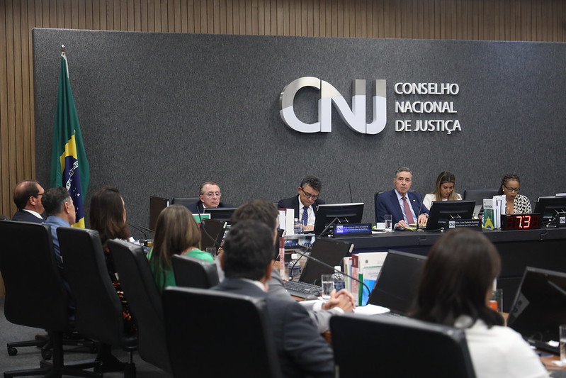 Plenário do Conselho Nacional de Justiça, sobre fundo de tom acinzentado a logo do CNJ, mesa em formato de “U”, composta pelo ministro Luís Roberto Barroso e os Conselheiros (as).