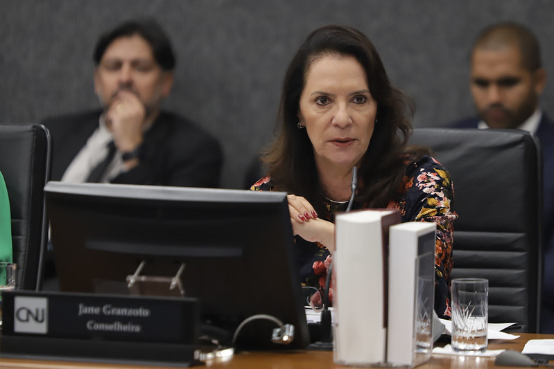 Imagem em plano médio da Conselheira Jane Granzoto, ela está sentada com as mãos apoiadas sobre a mesa, cabelos soltos de tom escuro e usando uma blusa de mangas longas de fundo escuro e estampa de florais.