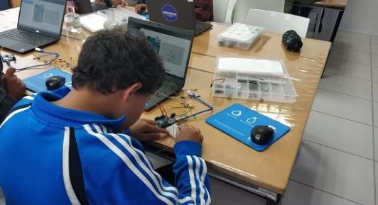 Imagem de um jovem sentado, manuseando fios elétricos, vestindo um moletom de tom azul. Sobre a mesa está alguns computadores e fios.