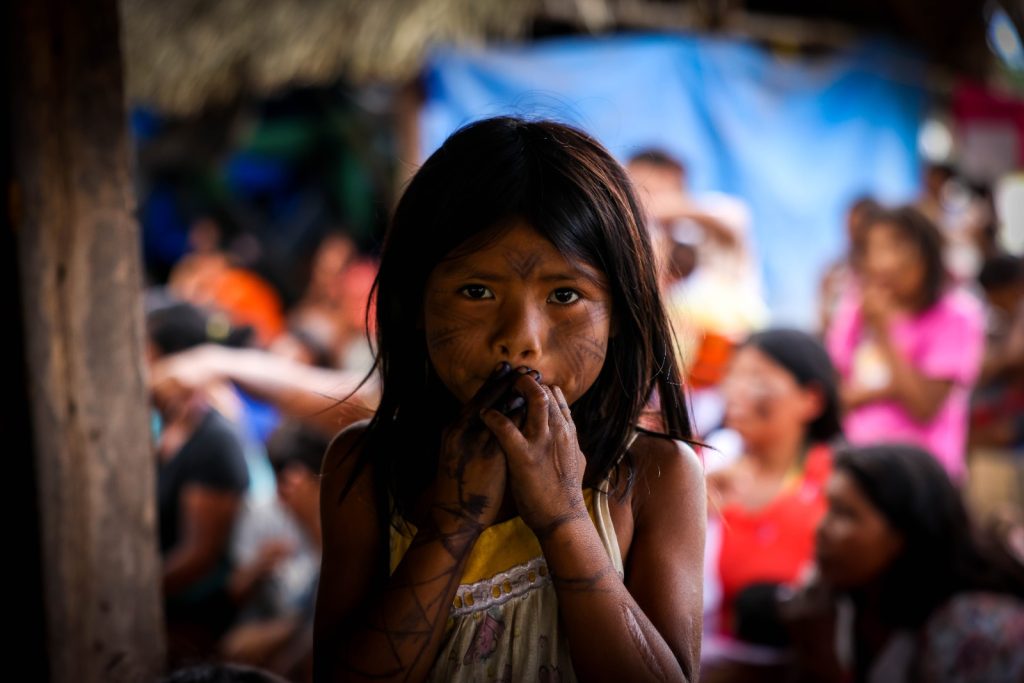 Fotografia de uma criança indígena, ela está de cabelos soltos, mãos na boca. Seu rosto, braços e mãos estão pintados com grafismos.