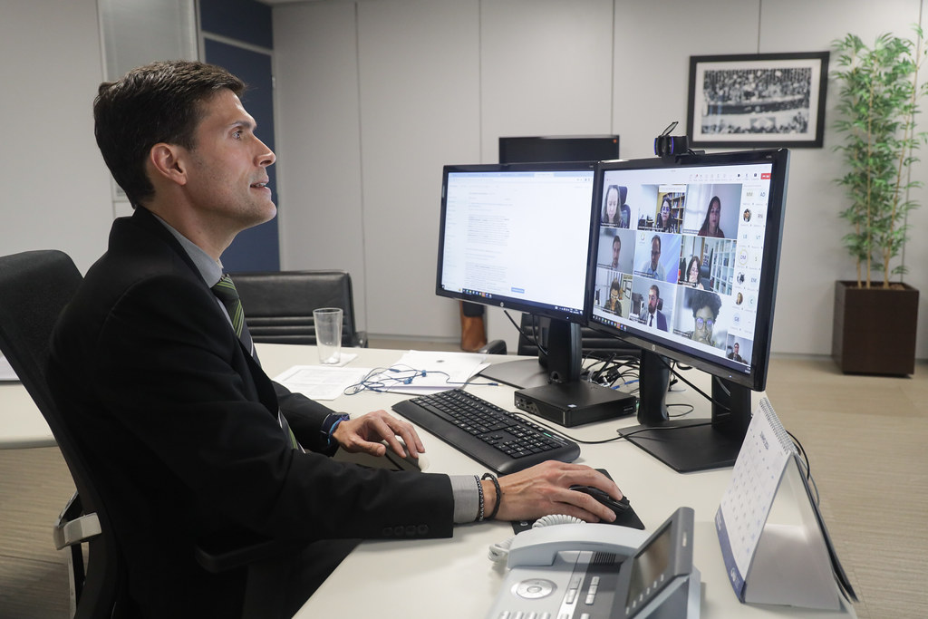 Fotografia em plano médio do Conselheiro do Conselho Nacional de Justiça, Pablo Coutinho Barreto, sentado, vestido formalmente e utilizando computador de mesa em um ambiente fechado. Sala de paredes brancas no canto inferior direito um jarro de planta e um quadro.