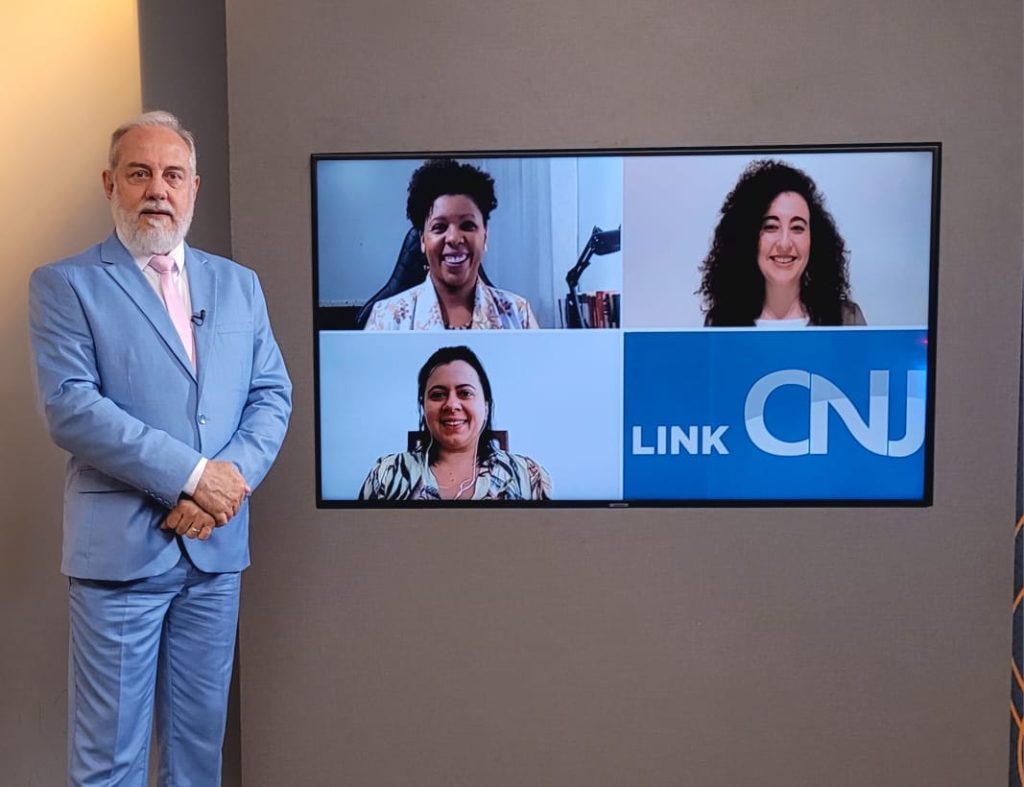 Foto do estúdio do programa com o apresentador posando ao lado do telão onde se vê as pessoas entrevistadas por videoconferência.