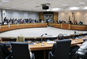 Fotografia em plano geral do Salão Pleno do Palácio da Justiça. Composta por mesa de formato circular em tom amadeirado, paredes, teto e piso em tons de branco. Autoridades sentados e vestidos formalmente. Ao fundo quadro artístico e bandeiras.