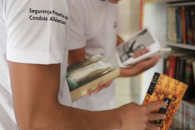 Sobre fundo desfocado, prateleira de livros, em primeiro plano duas pessoas fazendo leitura, vestidas com blusa branca, na manga da blusa está o texto: Segurança Protetiva via Conduta Alfabetizada.