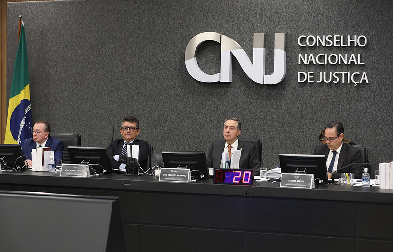 Fotografia em plano médio, plenário do Conselho Nacional de Justiça. Ao centro, na mesa em formato de U da esquerda para a direita estão o Presidente da OAB, Mansour Elias; Subprocurador-Geral da república, José Adônis Callou; Presidente do CNJ, Ministro Luís Roberto Barroso e o juiz auxiliar da presidência do CNJ, Gabriel Matos.