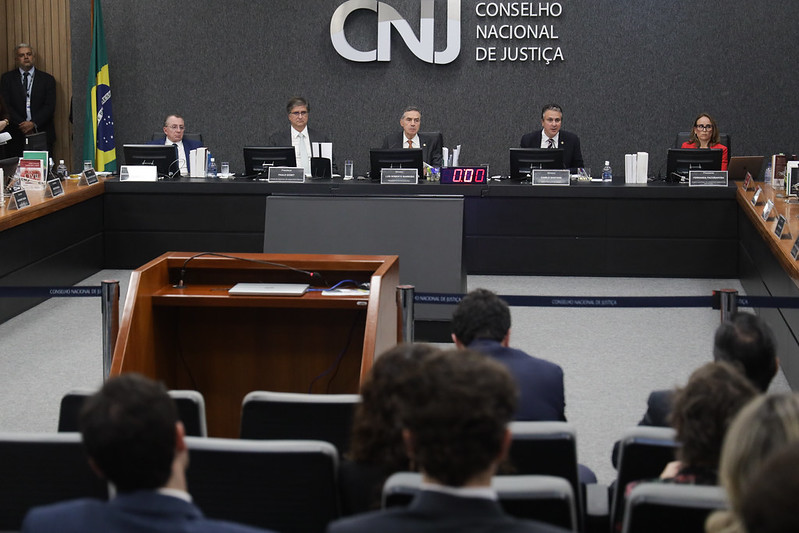 Fotografia em plano geral do plenário do Conselho Nacional de Justiça. Ao centro, na mesa em formato de U estão presentes ministros e demais autoridades. No fundo painel em tom de cinza e a logo do Conselho Nacional de Justiça (CNJ)