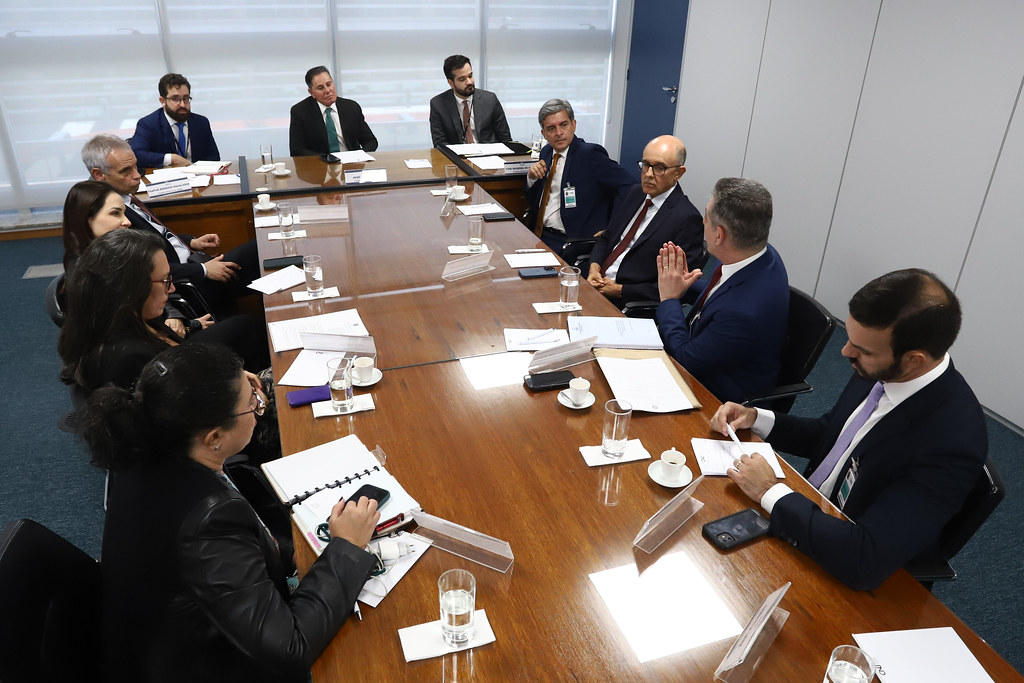 Sala de reunião composta por mesa em formato de T, homens e mulheres formalmente vestidos. Mesa de tom amadeirado e sala de paredes branca.