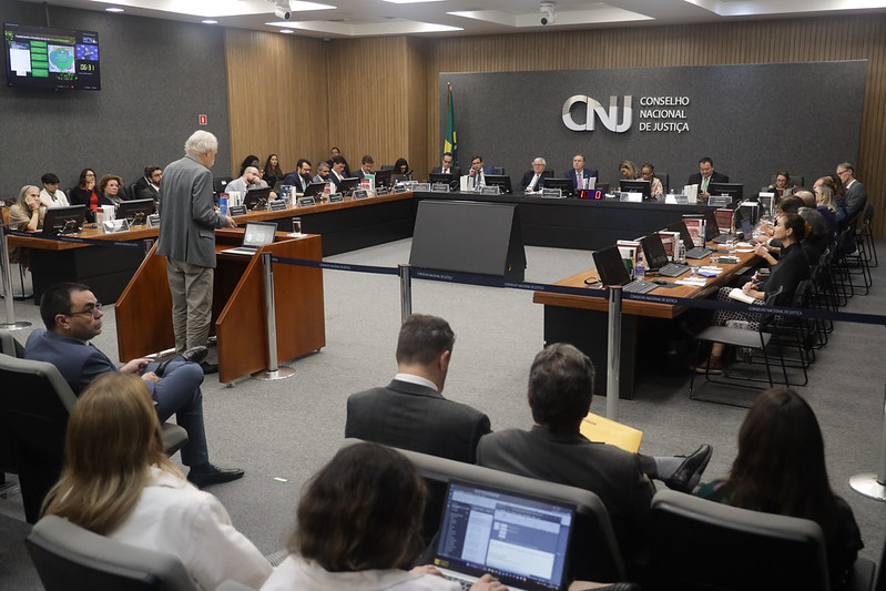 Fotografia em plano geral do plenário do Conselho Nacional de Justiça, em primeiro plano, plateia presente, em segundo plano, homem em pé em seu momento de fala e vestido formalmente. Ao fundo mesa em formato de U composta por autoridades do poder judiciário.