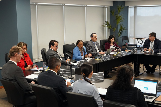 Sala de reunião composta por mesa de tom escuro em formato de U, paredes de tom azul, ao fundo uma planta ornamental. Presentes na mesa estão, conselheiros, juízes auxiliares do Conselho Nacional de Justiça e demais autoridades, todos vestidos formalmente.