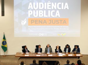 Fotografia em plano geral; em primeiro plano mesa composta por representantes do Conselho Nacional de Justiça e do Ministério da Justiça e Segurança Pública, vestidos formalmente. Ao fundo imagem projetada com texto; Audiência Pública - Pena Justa - Plano Nacional para o Enfrentamento do Estado de Coisas Inconstitucional nas Prisões Brasileiras, abaixo logo do CNJ e do Governo Federal.