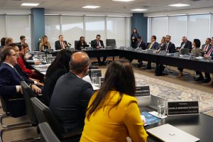 Sala de reunião composta por mesa de tom escuro em formato de “U”, na mesa está o presidente do CNJ, Ministro Luís Roberto Barroso e demais autoridades do poder judiciário, todos vestidos formalmente. A sala possui o teto branco, ao centro no chão, tapete florido.