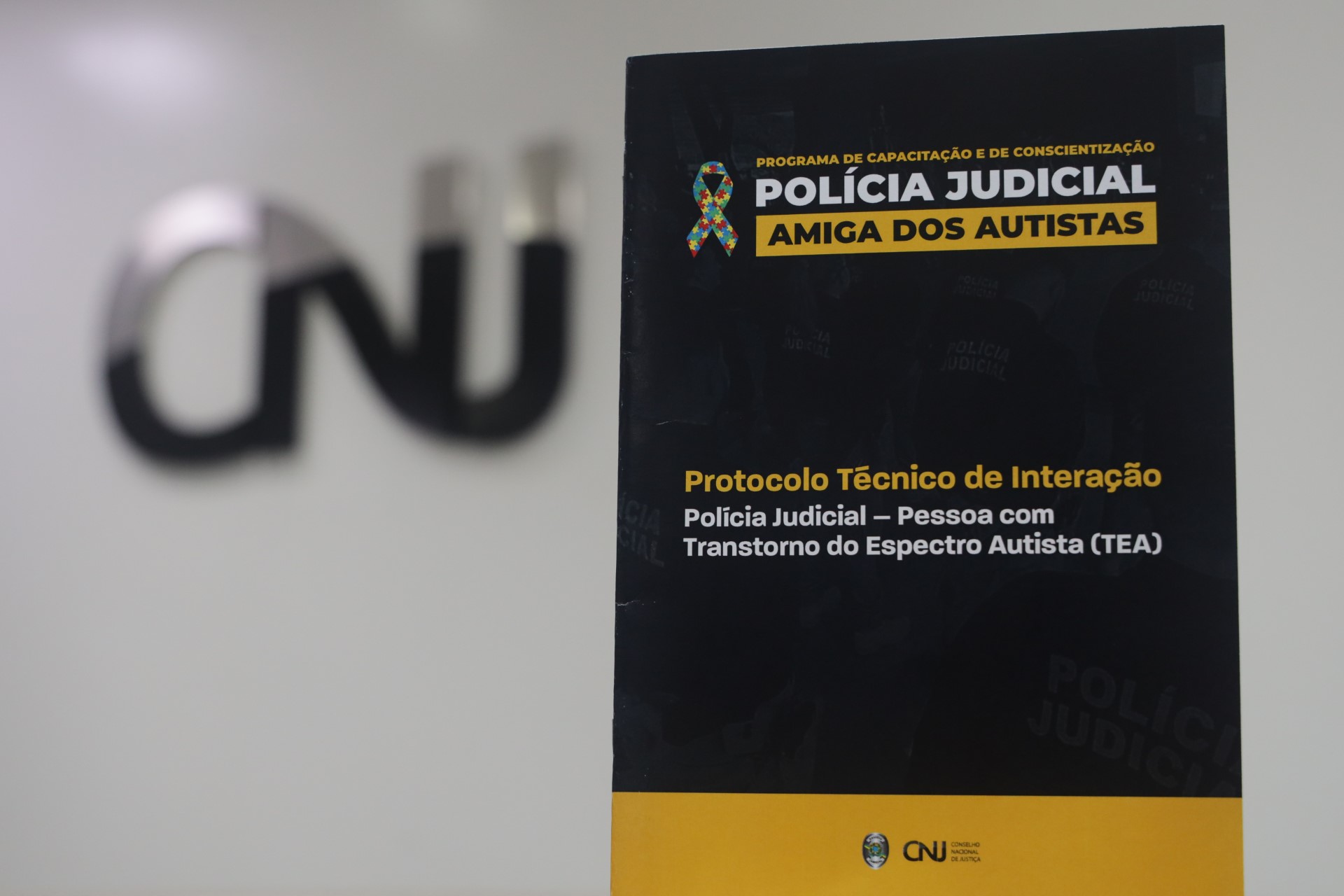 Panfleto com plano de fundo transparente contendo imagens de policiais judiciais, sobreposição em tons de preto e amarelo. No canto superior esquerdo o símbolo do autismo, centralizado o texto: Programa de Capacitação e de Conscientização - Policia Judicial - Amiga dos Autistas - Protocolo Técnico de Interação - Polícia Judicial - Pessoa com Transtorno do Espectro Autista (TEA).