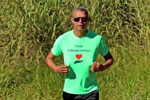 Fotografia de um homem usando óculos de sol, vestindo short de tom escuro e blusa de tom verde, na blusa está a frase - Treinão A vida pode continuar, abaixo ilustração de uma mão e coração.