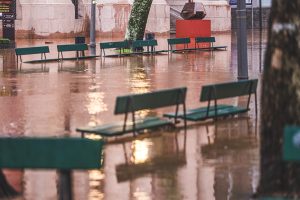 Fotografia de uma praça em Porto Alegre alagada. Há bancos de cor verde, uma árvore e dois postes de luz. Ao fundo, há uma escada. 