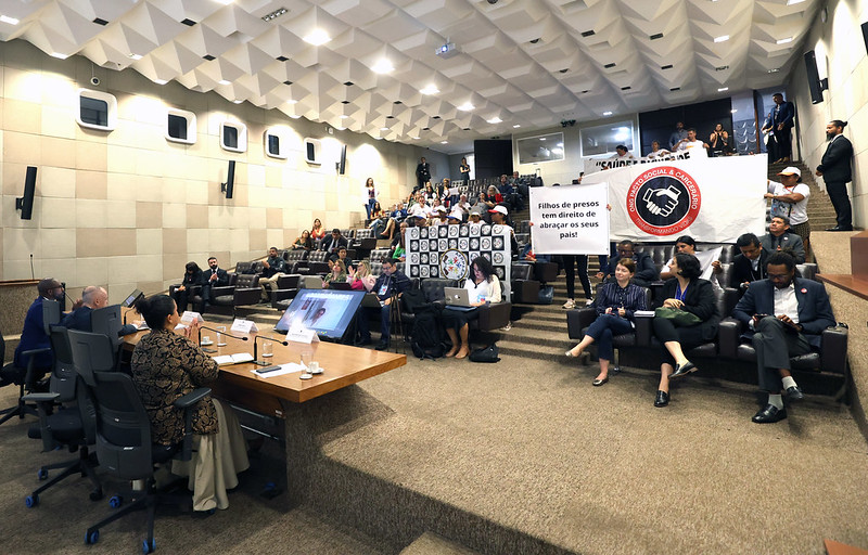 Fotografia em plano geral do auditório do Ministério da Justiça. Em primeiro plano mesa composta por integrantes da Audiência Pública, todos vestidos formalmente. Ao fundo, plateia participante, usando computadores e cartazes em manifesto.