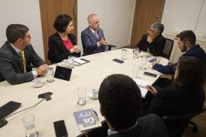 Sala de reunião de portas em tom amadeirado e paredes brancas, ao centro, mesa de tom claro composta por seis pessoas, homens e mulheres vestidos formalmente. Sobre a mesa estão copos com água, xícaras de café, agendas e celulares.