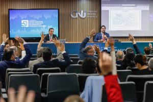 Fotografia do auditório do Conselho Nacional de Justiça durante o encerramento do 2° Congresso do Fórum Nacional de Recuperação Empresarial e Falências (Fonaref). Em primeiro plano, plateia de mãos levantadas, em segundo plano, mesa composta pelo conselheiro Marcos Vinicius Jardim e o advogado Daniel Carnio, ambos vestidos formalmente. Ao fundo painel de tom amadeirado com a logo do CNJ e telas de transmissão