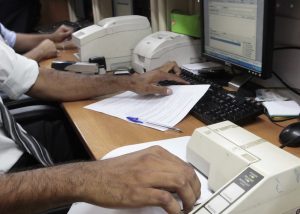 Fotografia em plano médio, mesa de tom amadeirado composta por computador de mesa, impressoras e materiais de escritórios.