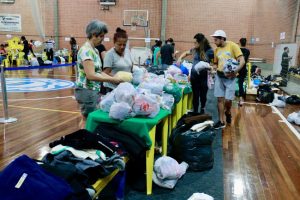 Fotografia de uma quadra de esportes sendo utilizada como centro de atendimento e acolhimento para as vítimas das enchentes em Porto Alegre. Quadra cheia com pessoas, mesas de plásticos, sacolões e roupas.